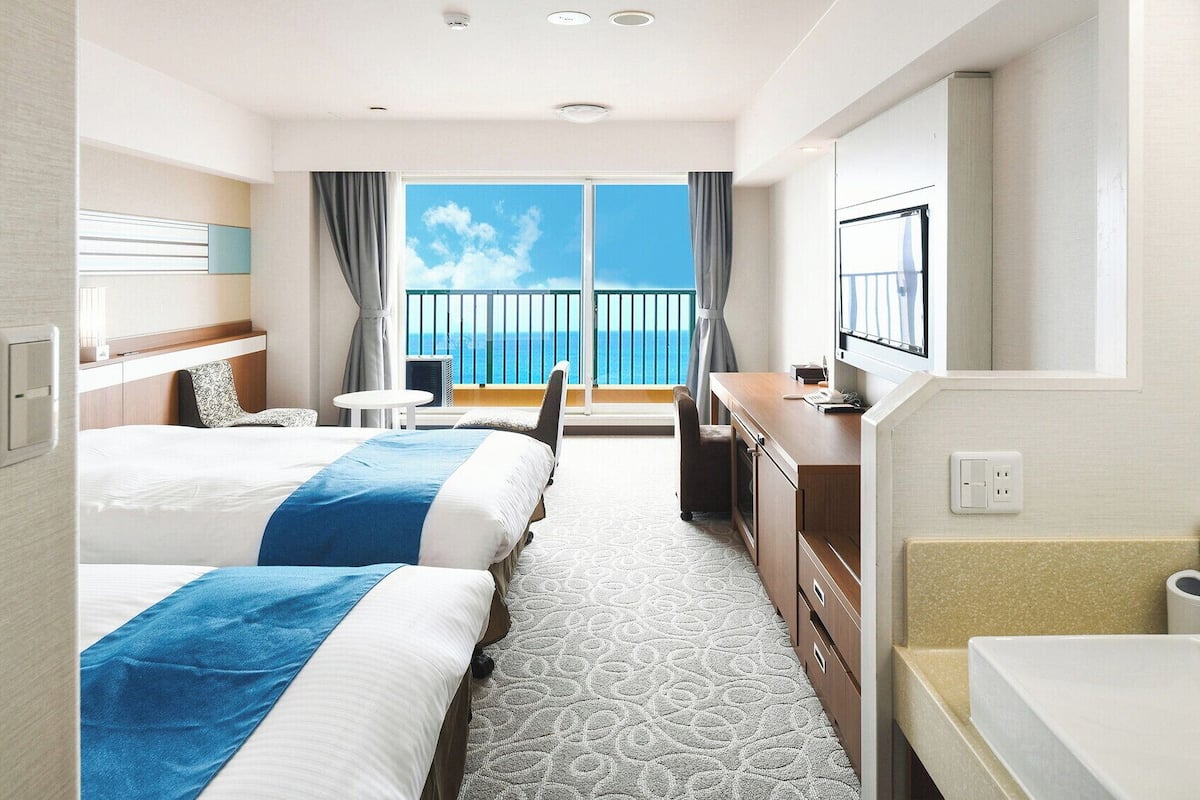 Une chambre d’hôtel avec deux lits, un bureau, une chaise, une télévision et un balcon donnant sur la mer.