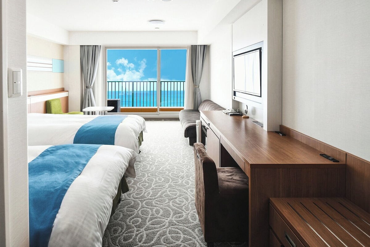 Une chambre d’hôtel avec deux lits, un bureau, une chaise, une petite table et une grande fenêtre donnant sur la mer.