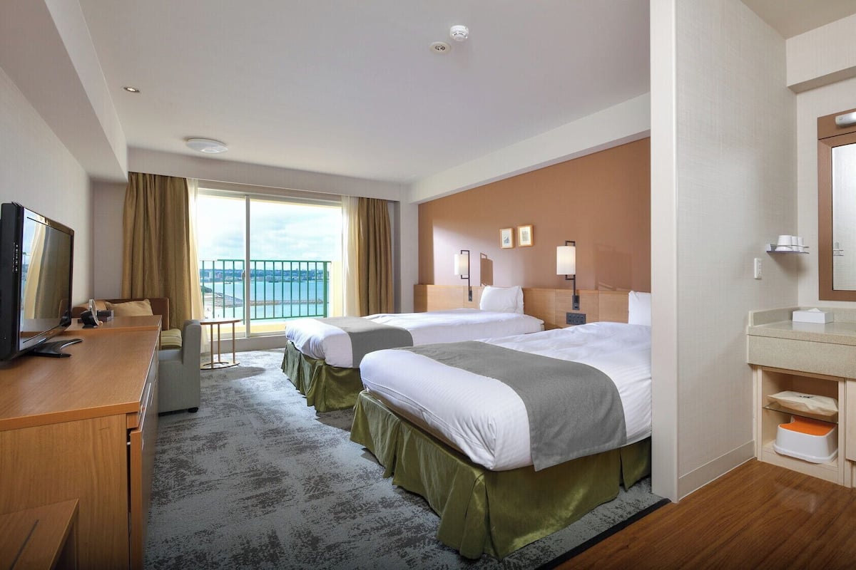 Une chambre d’hôtel avec deux lits, un bureau avec une télévision, une salle de bain avec un miroir et un lavabo, et un balcon avec vue.