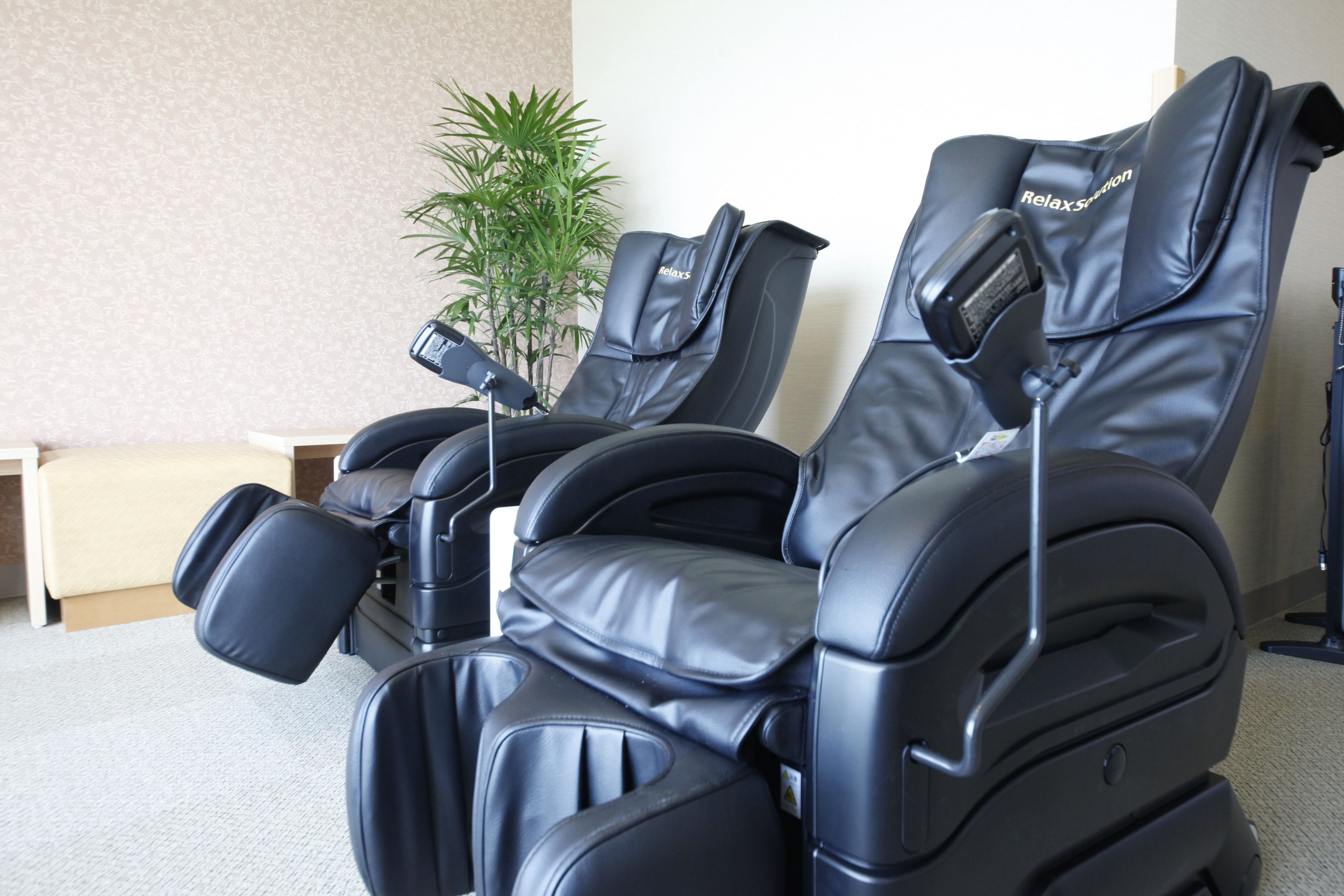 A massage chair with a control panel, positioned in a room with a plant and a small table.