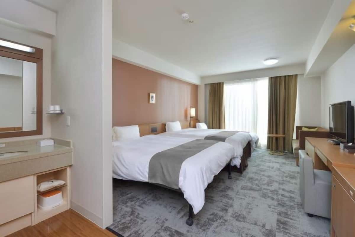 Une chambre d’hôtel avec deux lits, un bureau, une télévision, une salle de bain avec un miroir et une fenêtre avec des rideaux.