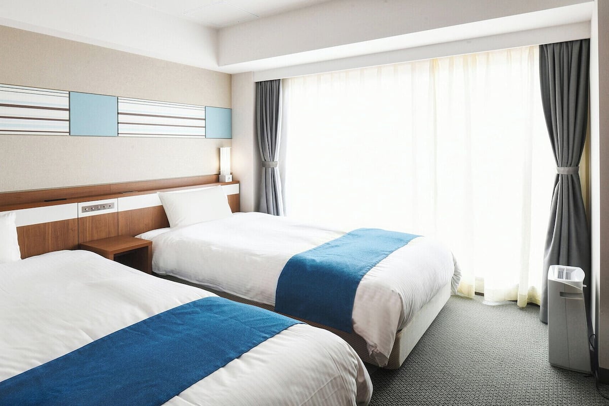 Une chambre d’hôtel avec deux lits, une tête de lit en bois, une table de chevet, une lampe fixée au mur et une grande fenêtre avec des rideaux.