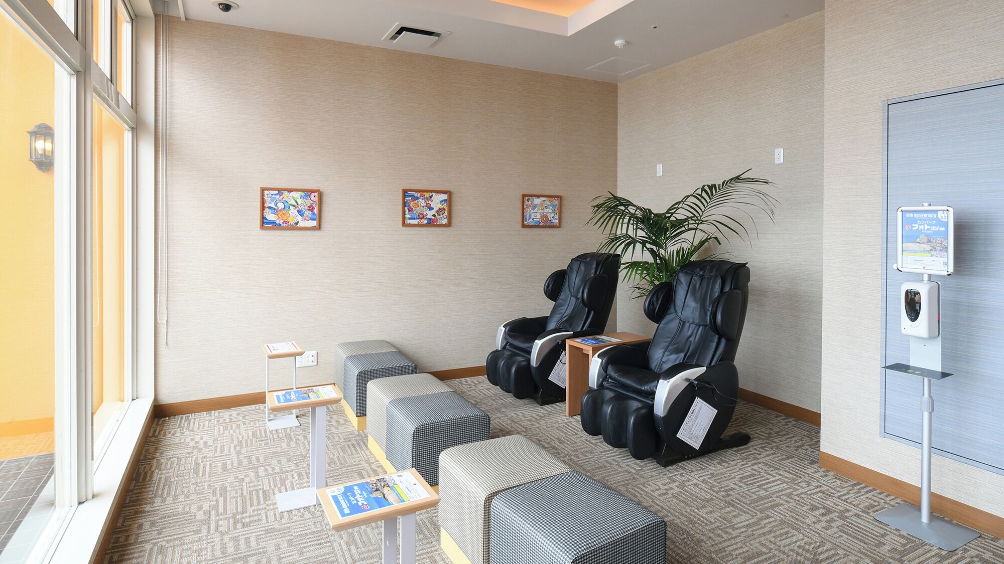 Uma área de recepção de hotel com espaço para sentar, cadeiras de massagem, balcão de recepção e um quadro de informações fixado na parede.