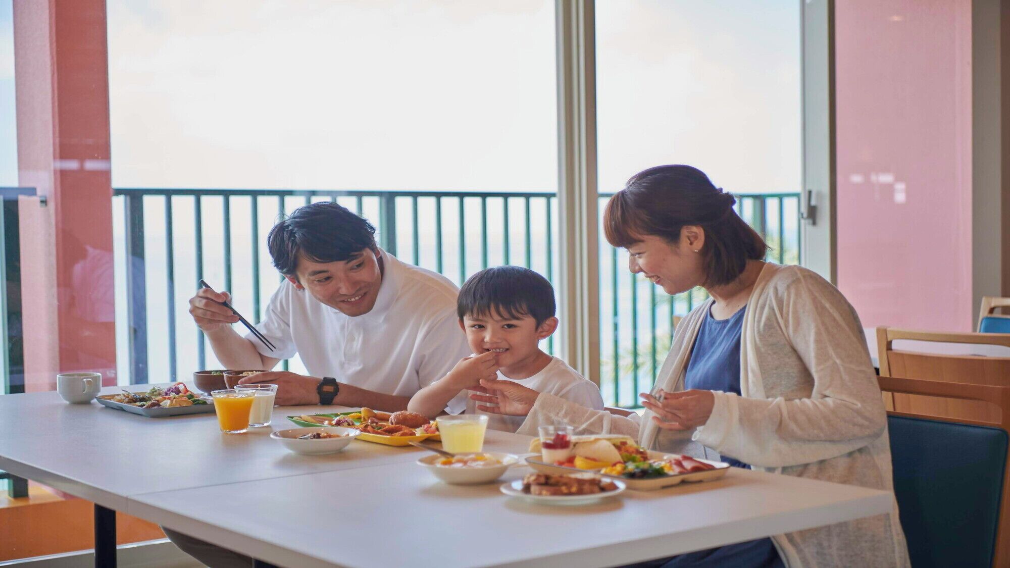 Una mesa de comedor con diversos platos y bebidas, una ventana con persianas y una barandilla de balcón.