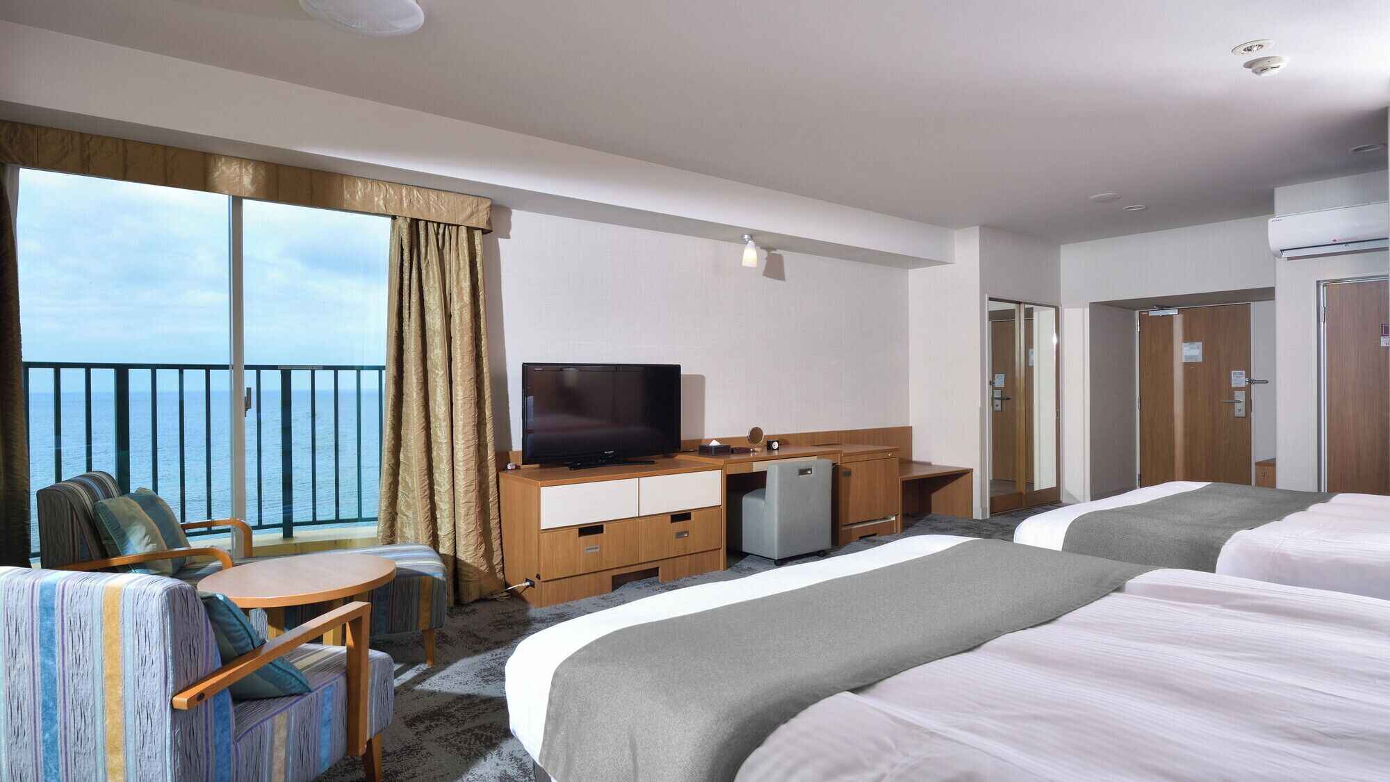 Habitación de hotel con dos camas, un escritorio con televisor, un refrigerador pequeño y un balcón con vista al mar.