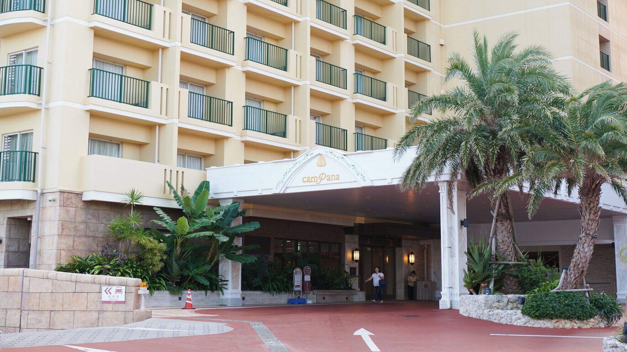 A hotel entrance with a covered walkway, palm trees, and a sign reading "Camana".