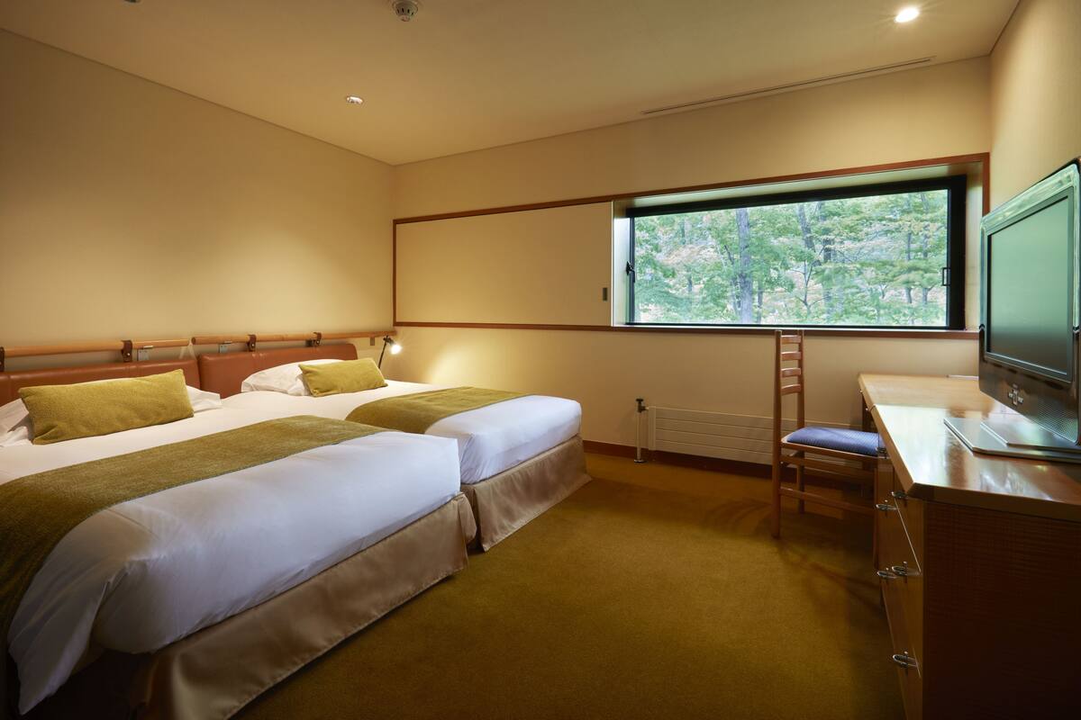 Een hotelkamer met twee bedden, een bureau met een computer, een raam met uitzicht op bomen, en een radiator.