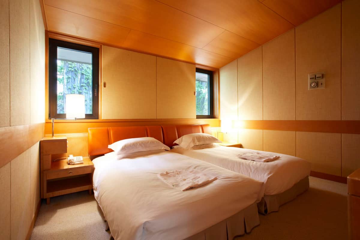 Een tweepersoonskamer met twee bedden, een houten hoofdeinde, een nachtkastje, een lamp en een raam met uitzicht op bomen.