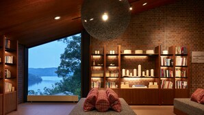 Un coin lecture confortable, avec une grande fenêtre donnant sur un lac, entouré d’étagères remplies de livres et d’objets décoratifs.