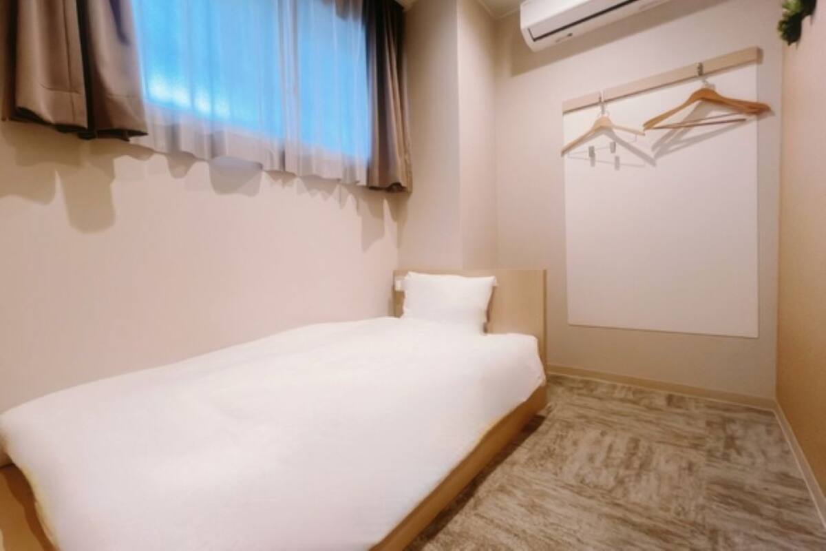 Un lit simple avec un matelas et une tête de lit blancs, une fenêtre avec des rideaux légers, un appareil de climatisation et une armoire murale avec des cintres en bois.
