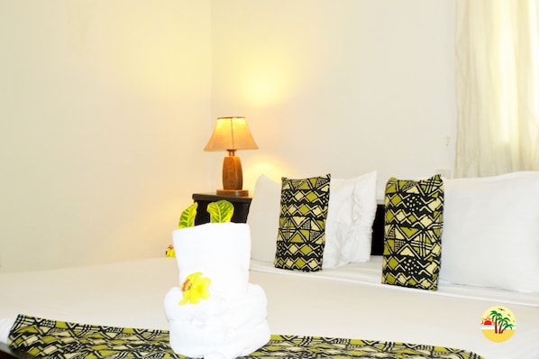 A hotel room with a bed, a nightstand with a lamp, patterned pillows, and a towel with a flower.