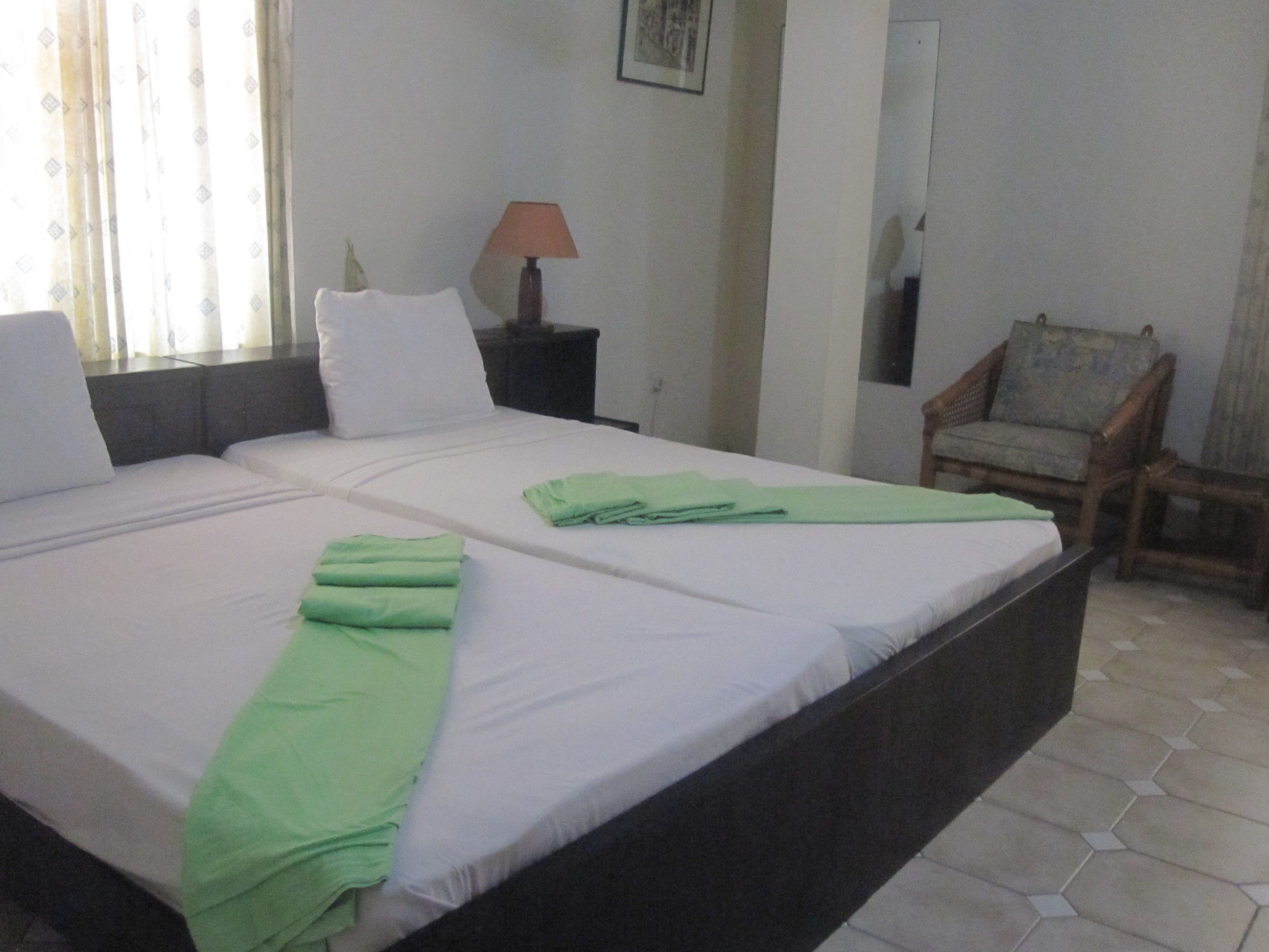 Cama doble con sábanas blancas y toallas verdes, mesita de noche con lámpara, una silla y ventana con cortinas.