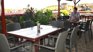 An outdoor cafe with wooden tables and wicker chairs, surrounded by greenery.