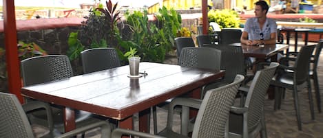An outdoor cafe with wooden tables and wicker chairs, surrounded by greenery.