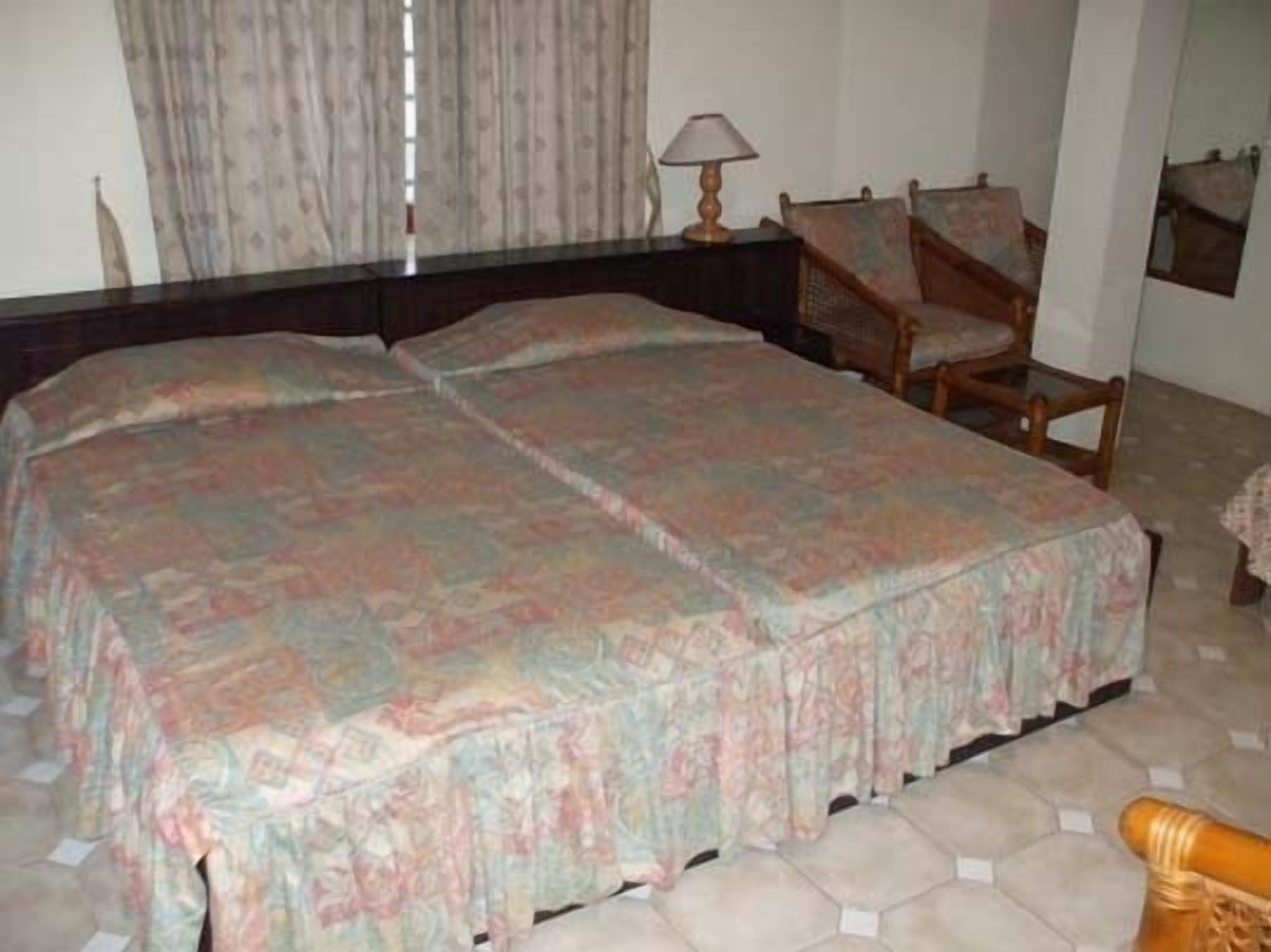 Quarto com cama grande, cadeira de madeira, mesa de cabeceira com abajur e janela com cortinas.