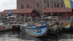 A stone building with a red-tiled roof, several boats docked in front, and a flag with blue and yellow colors.