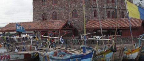 A stone building with a red-tiled roof, several boats docked in front, and a flag with blue and yellow colors.