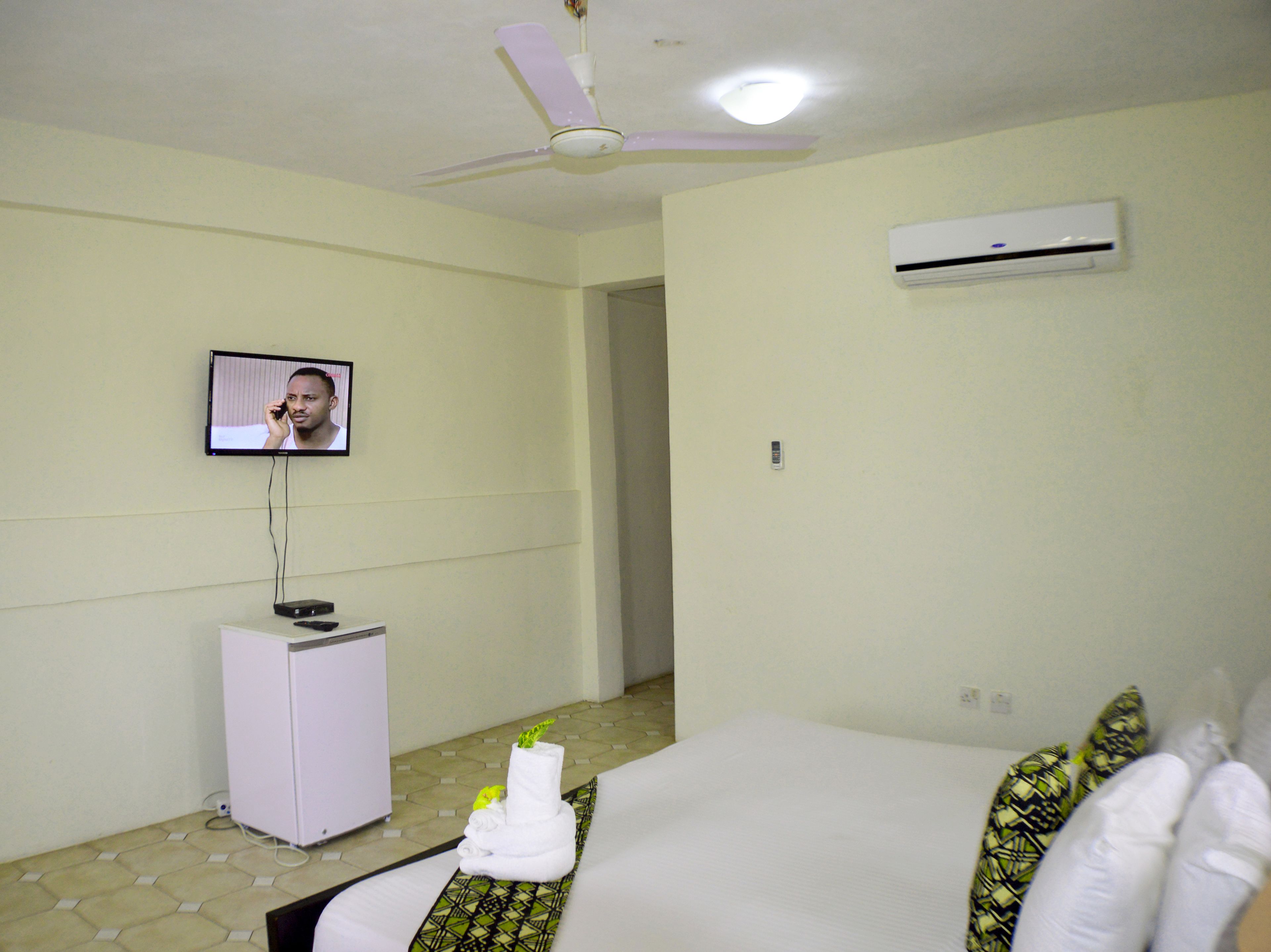 Una habitación de hotel con cama, televisor montado en la pared, un pequeño mueble blanco y un equipo de aire acondicionado.