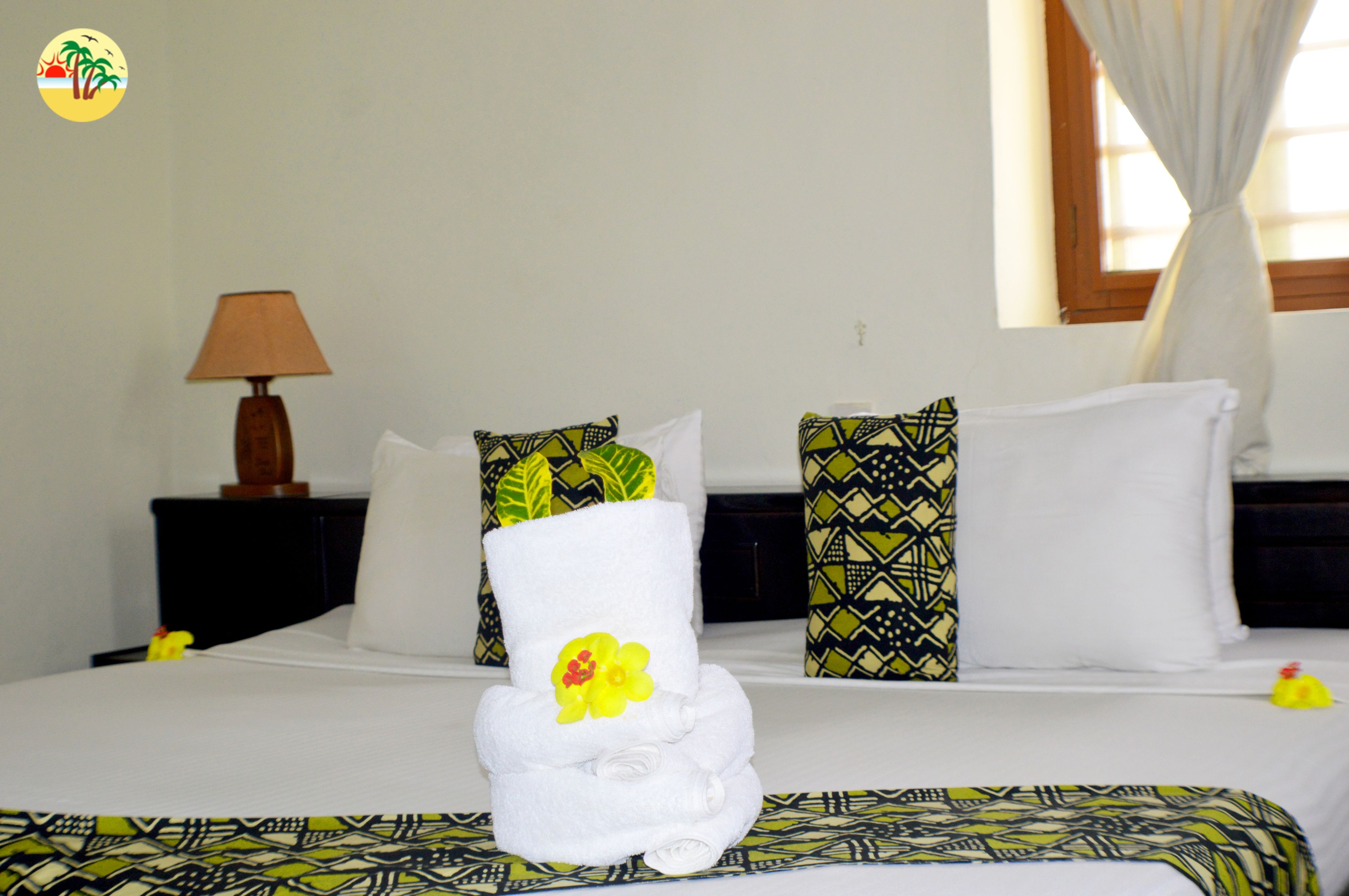 A neatly made bed with a towel centerpiece, patterned pillows, a nightstand with a lamp, and a window with curtains.