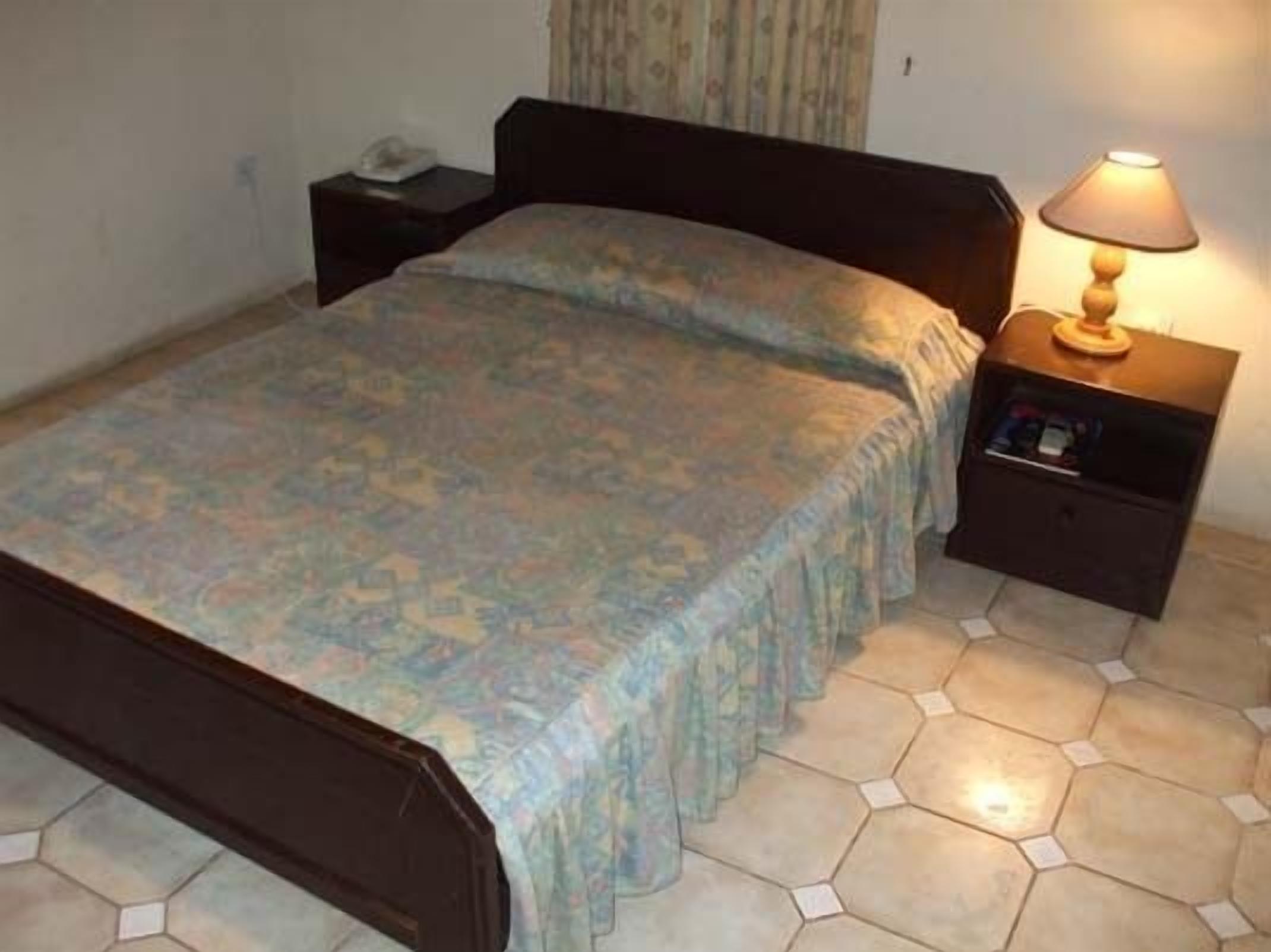 Una cama individual con una colcha estampada, una mesita de noche con lámpara y suelo de baldosas.