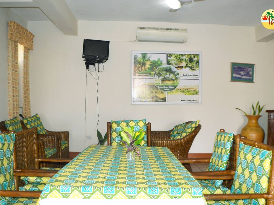 A dining area with a table covered in a patterned tablecloth, surrounded by wicker chairs with cushions. A television is mounted on the wall, and there is a framed picture with text and images.