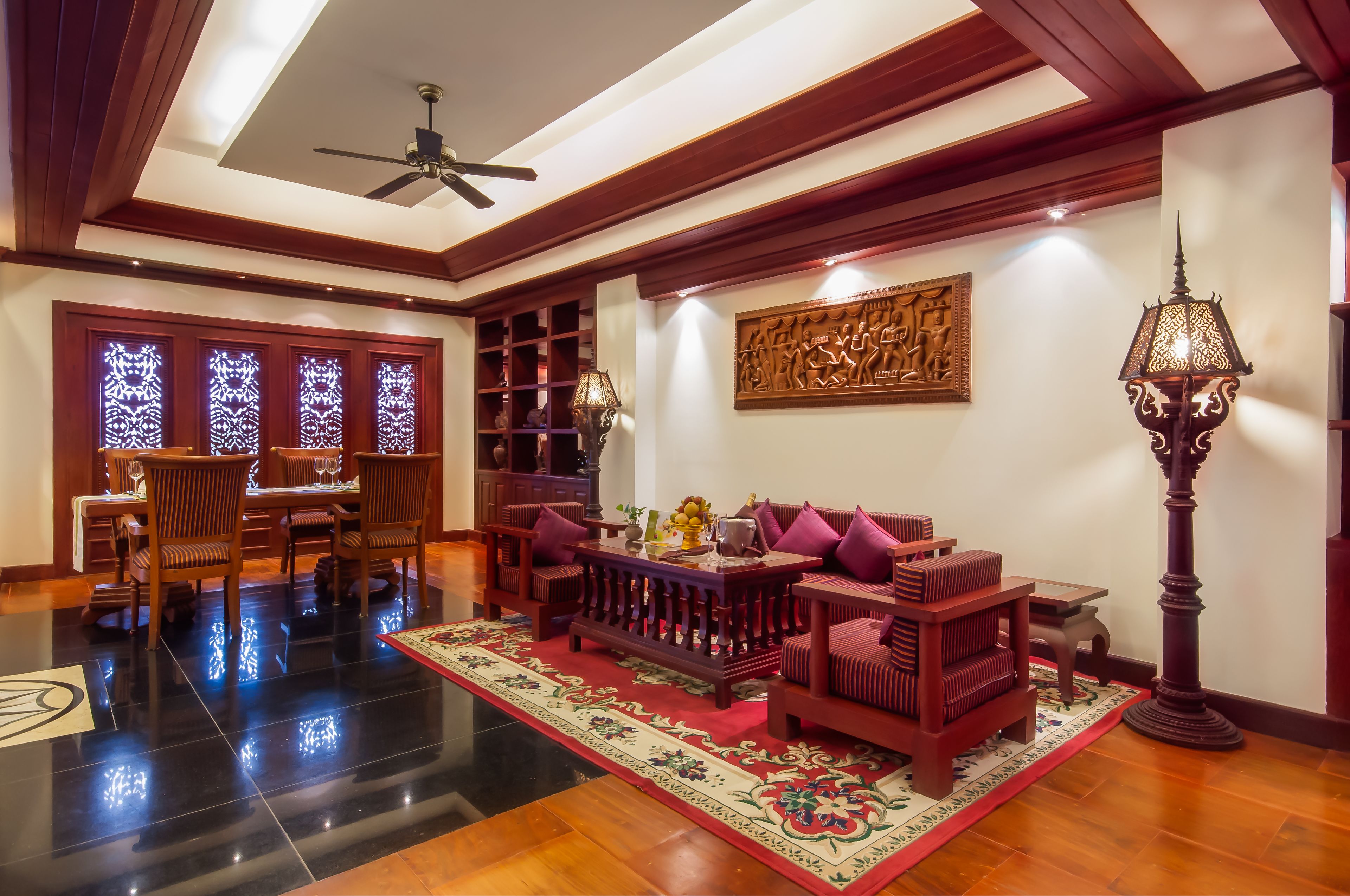 A spacious living area with a wooden ceiling, a ceiling fan, a dining table with chairs, a sofa with cushions, a patterned rug, and a tall floor lamp.
