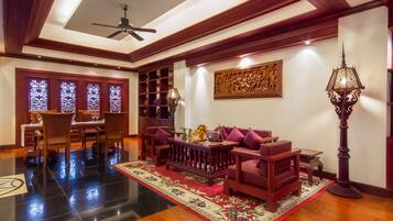 A spacious living area with a wooden ceiling, a ceiling fan, a dining table with chairs, a sofa with cushions, a patterned rug, and a tall floor lamp.