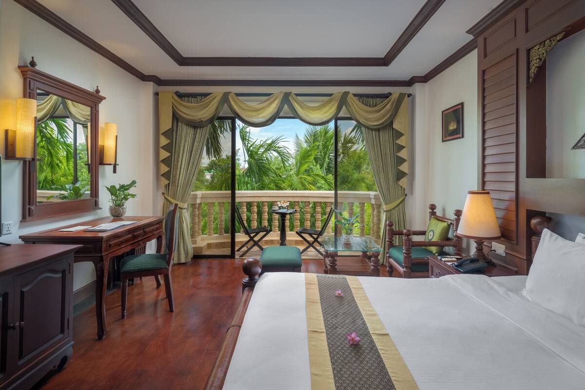 Ein Hotelzimmer mit einem großen Bett, einem Schreibtisch mit Stuhl, einem kleinen Tisch mit Stühlen und einem Balkon mit Vorhängen.