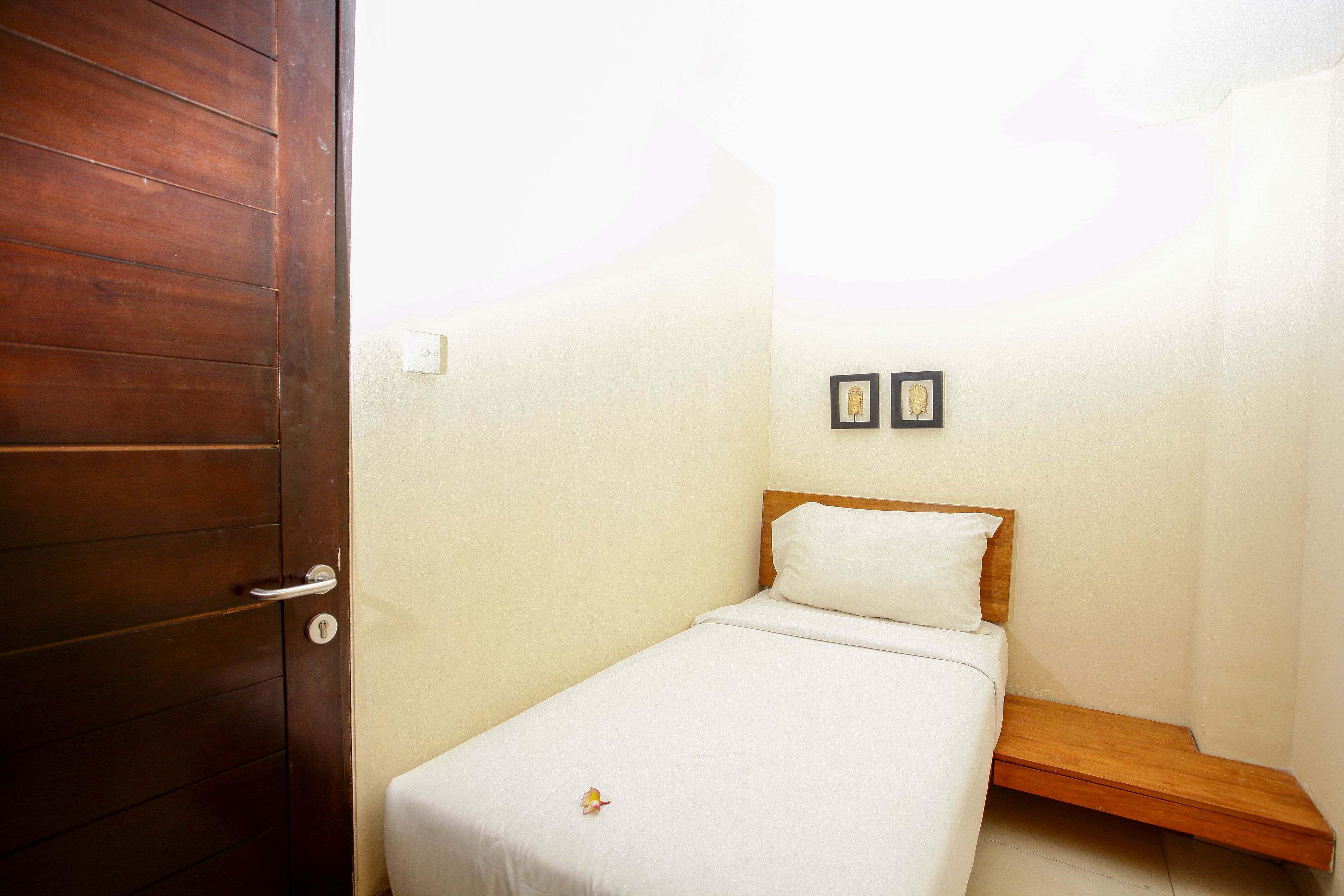 Une petite chambre avec un lit simple, une porte en bois, un lit blanc, une petite étagère en bois et deux tableaux encadrés accrochés au mur.