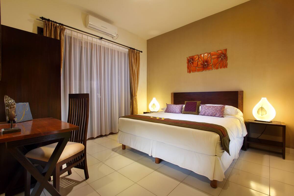 A hotel room with a bed, a desk, and a chair. There is a window with curtains, a wall-mounted air conditioner, and a decorative wall piece.
