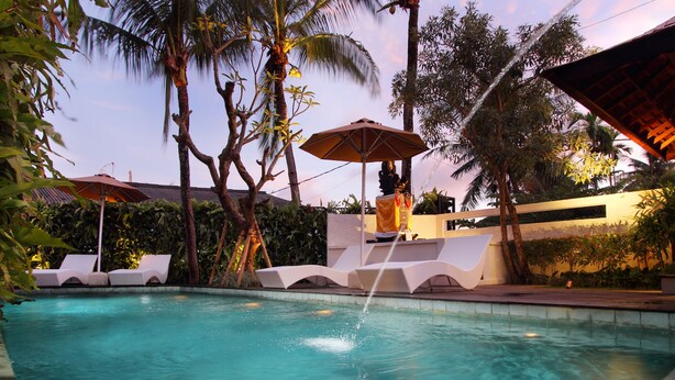 A swimming pool with lounge chairs and an umbrella, surrounded by palm trees and a building with a white facade. - Berawa Beach Residence (Tibubeneng)