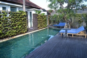 A swimming pool with a wooden deck, two blue lounge chairs, and a small table.