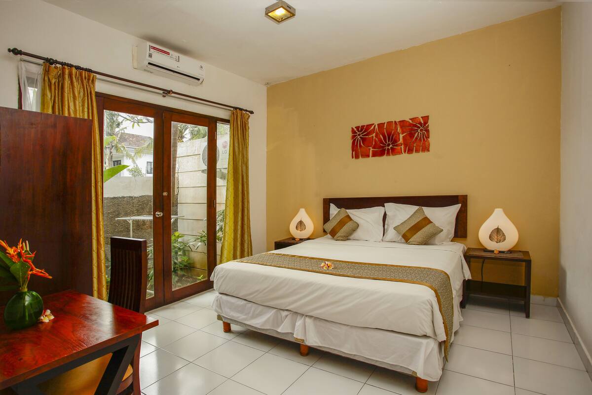 A hotel room with a bed, a desk, a chair, a nightstand, a lamp, and a wall decoration.