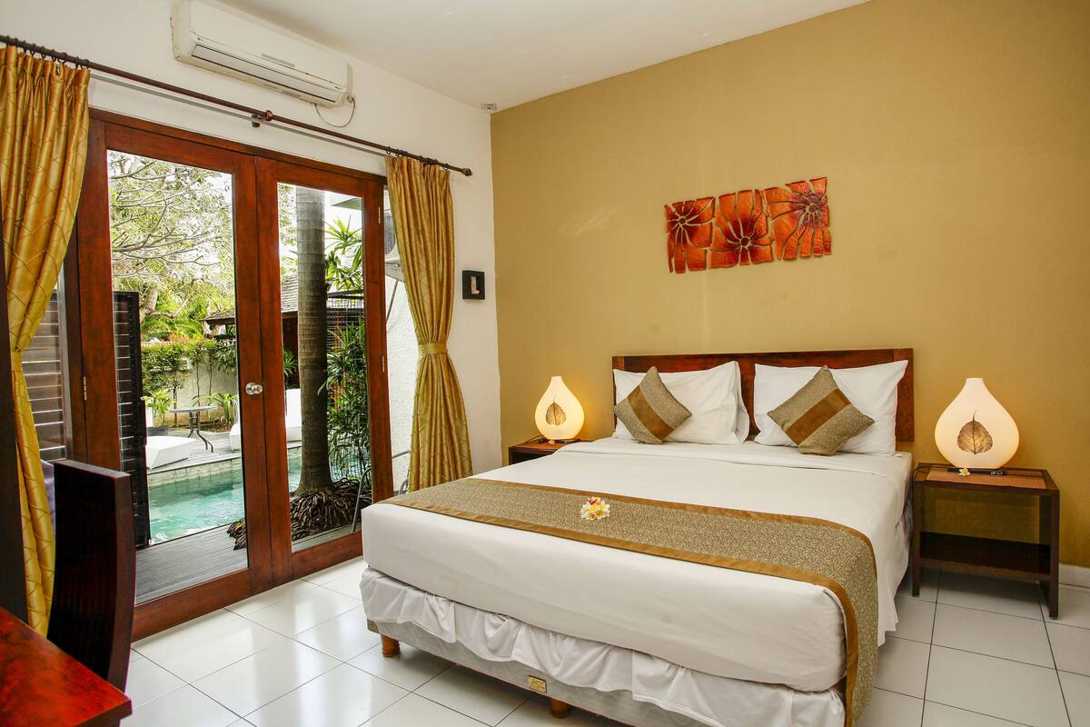A hotel room with a large bed, two bedside tables with lamps, a view of a pool through a sliding door, and a tropical-themed wall decoration.