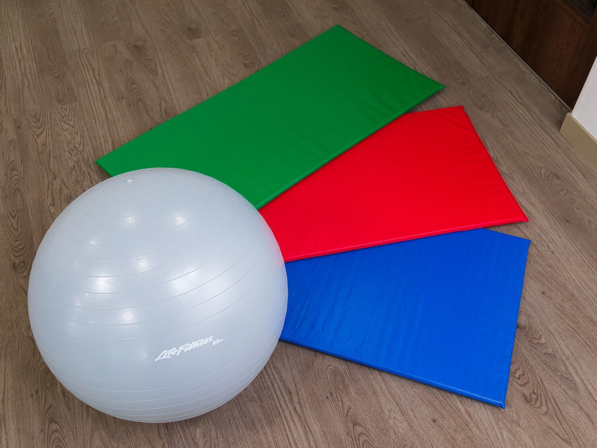 Una pelota de ejercicios blanca con la etiqueta 'Life Fitness' sobre un piso de madera, con cuatro colchonetas de colores (verde, rojo, azul y amarillo) apiladas encima.