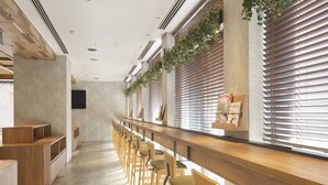 Une longue pièce étroite, avec des bancs et des chaises en bois disposés le long des murs, de grandes fenêtres équipées de stores et un sol en béton.