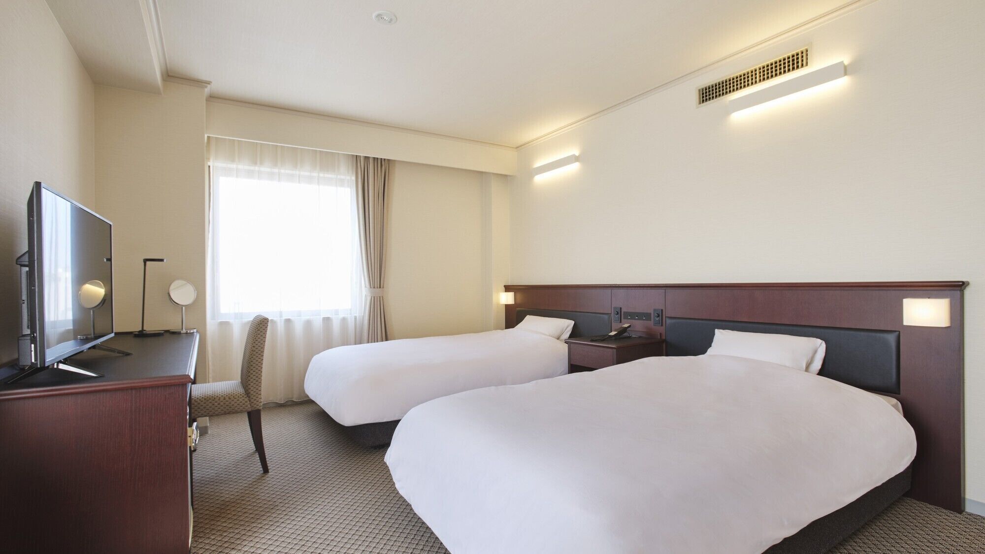 Ein Hotelzimmer mit zwei Betten, einem Schreibtisch mit Fernseher, einem Sessel, einem Fenster mit Vorhängen und einer Wandlampe.
