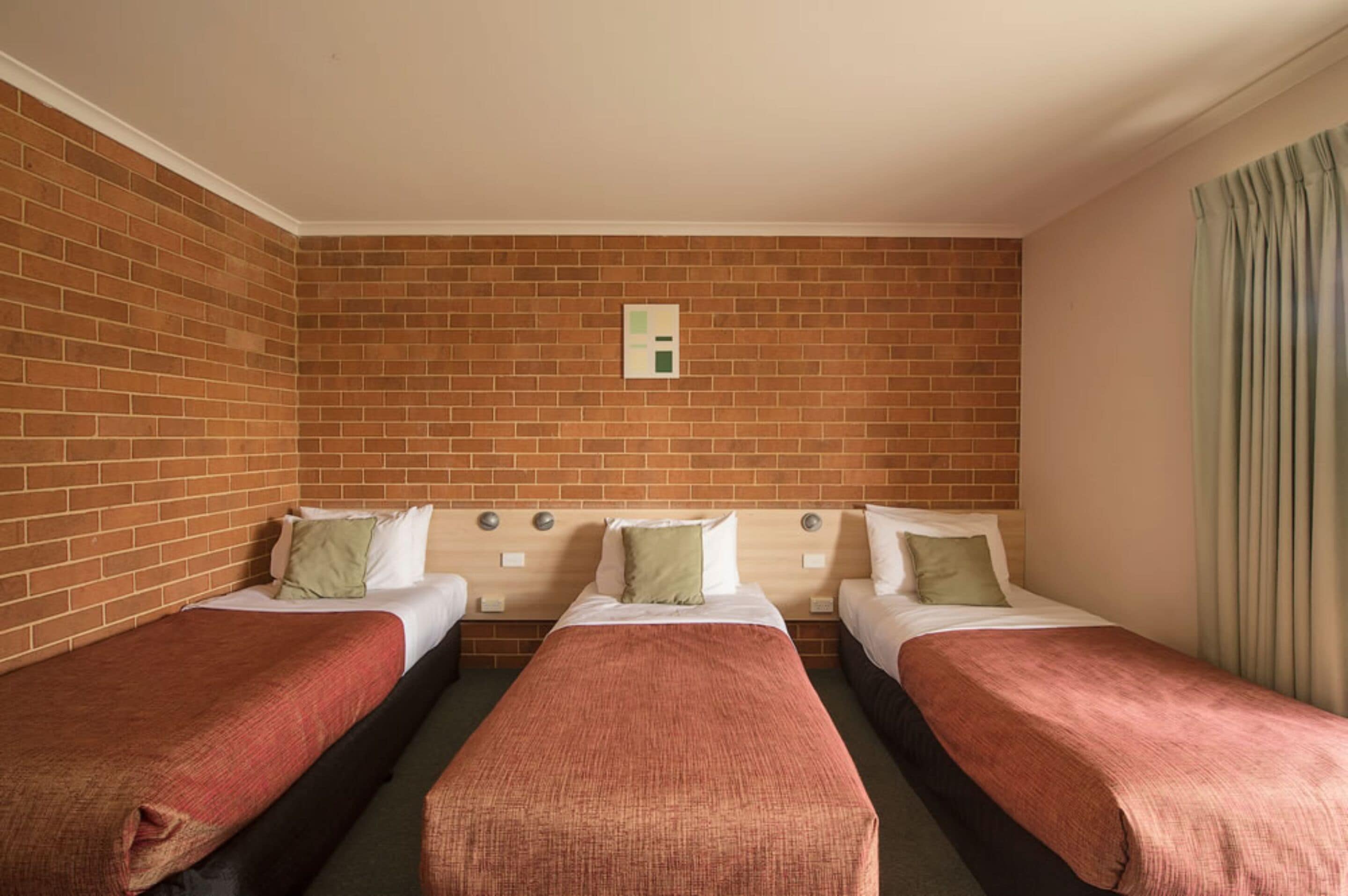 Ett hotellrum med tre enkelsängar, en tegelvägg och ett fönster med gardiner.