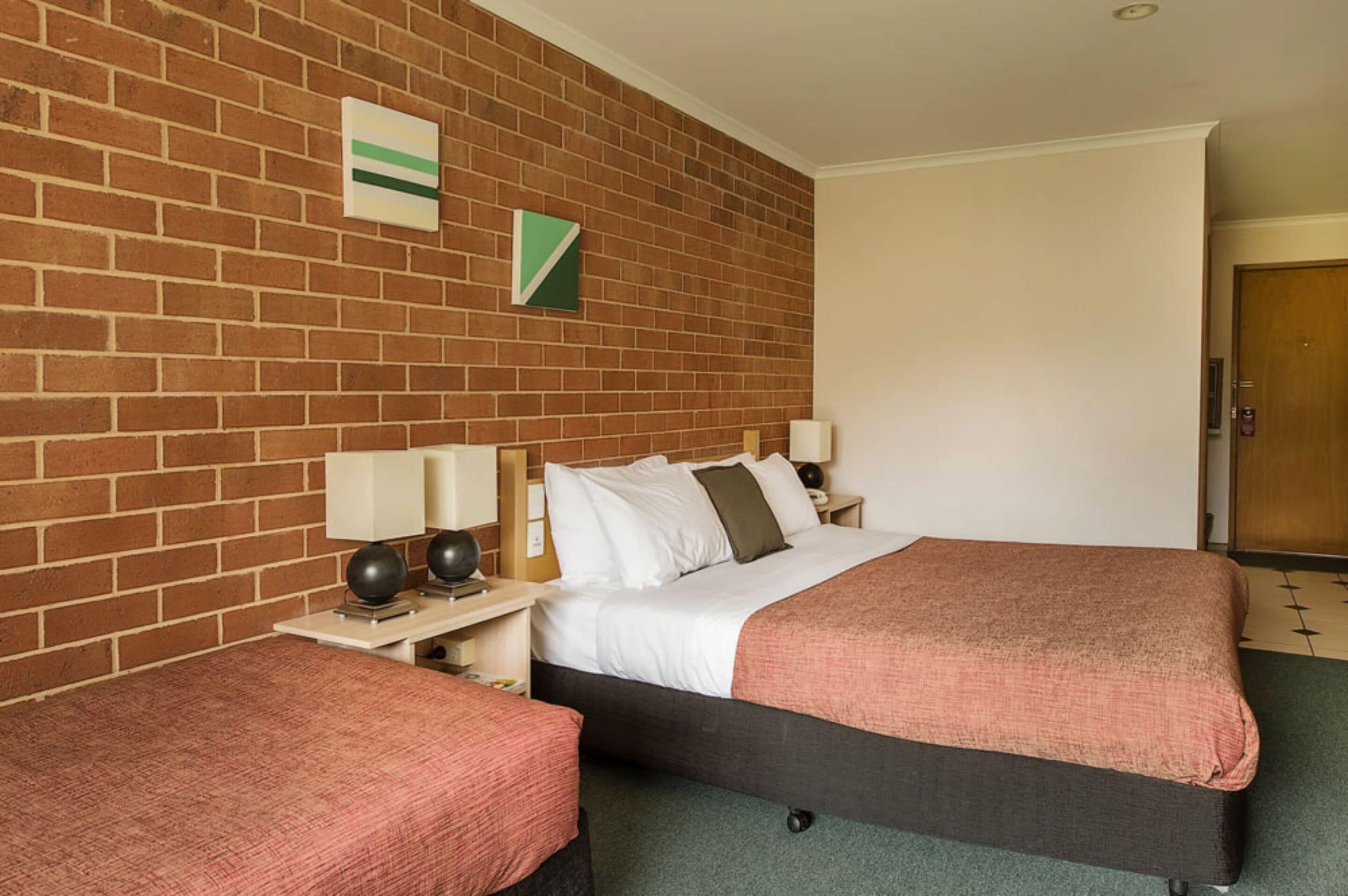 A hotel room with a brick wall, a bed with a dark frame, a bedside table with a lamp, and a small table with a chair.