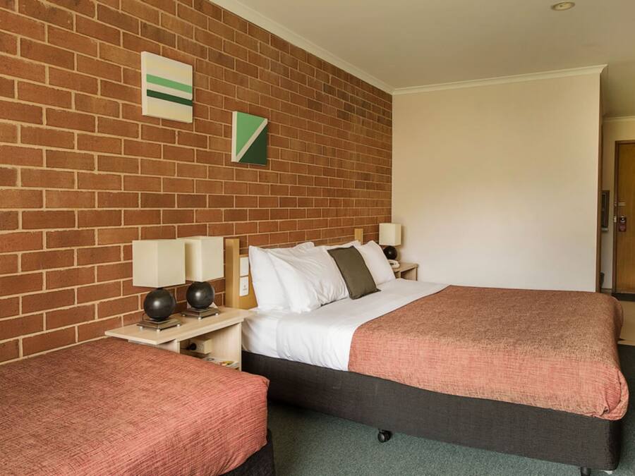 A hotel room with a brick wall, a bed with a dark frame, a bedside table with a lamp, and a small table with a chair.