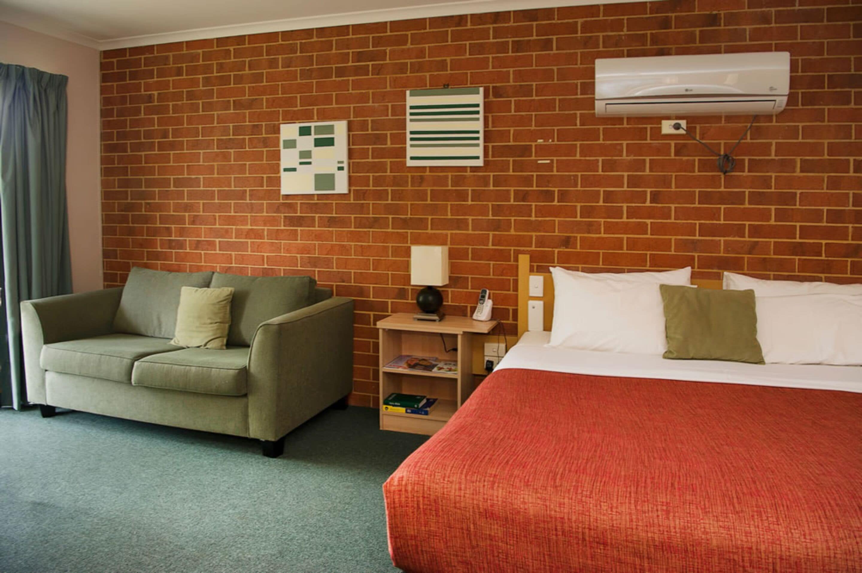 Una habitación de hotel con pared de ladrillo, una cama con almohadas blancas y verdes, un sofá beige, una mesita de noche con lámpara y un aire acondicionado instalado en la pared.
