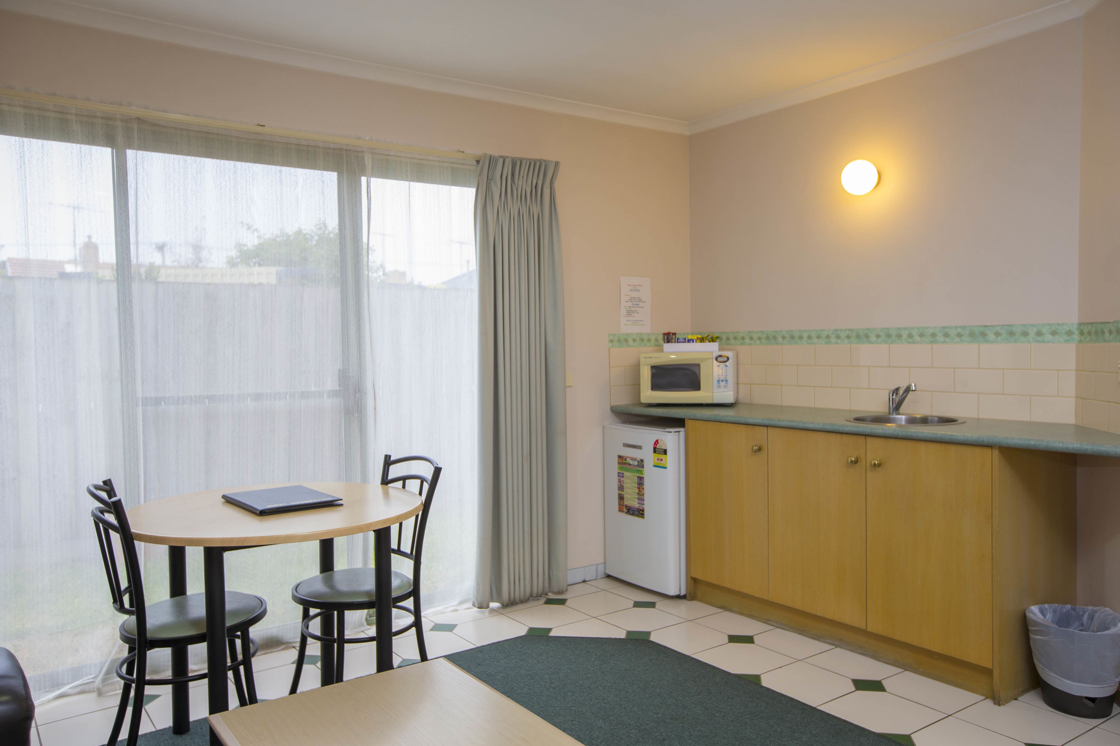Una cocina pequeña con microondas, lavamanos y refrigerador. Hay una mesa de comedor con sillas y una ventana con cortinas.