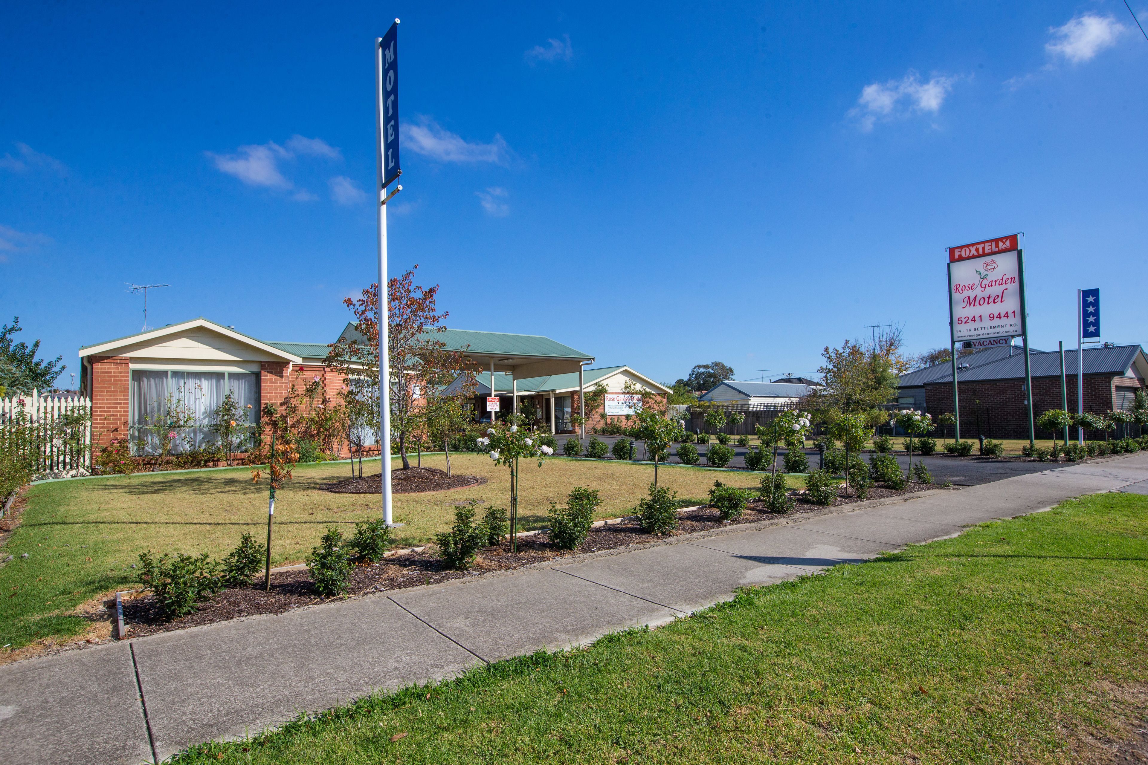A Rose Garden Motel with a clear blue sky, a well-maintained lawn, and a parking area.
