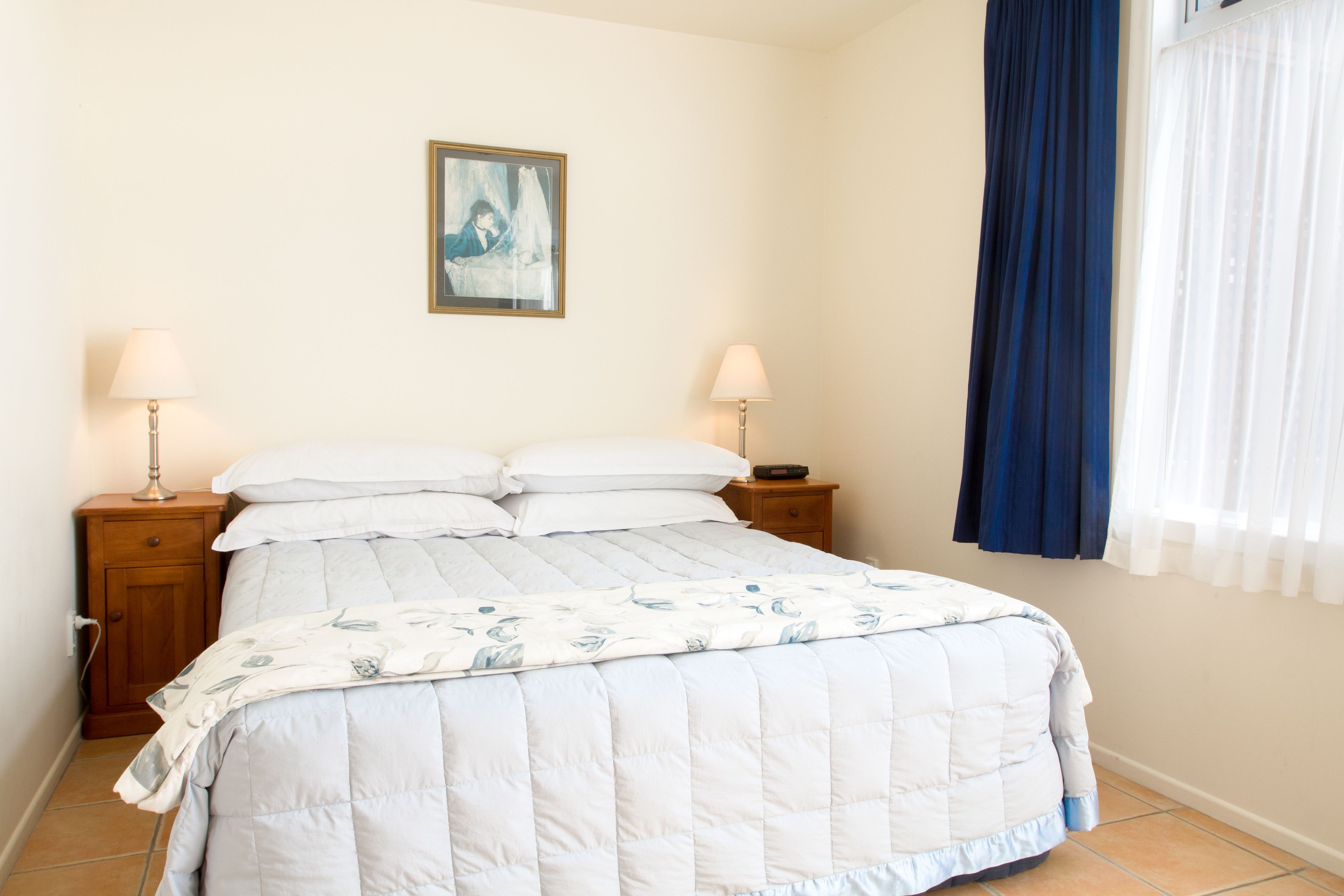 Una cama bien hecha con colcha blanca y azul, dos lámparas de noche, una mesita de madera pequeña y un cuadro en la pared.