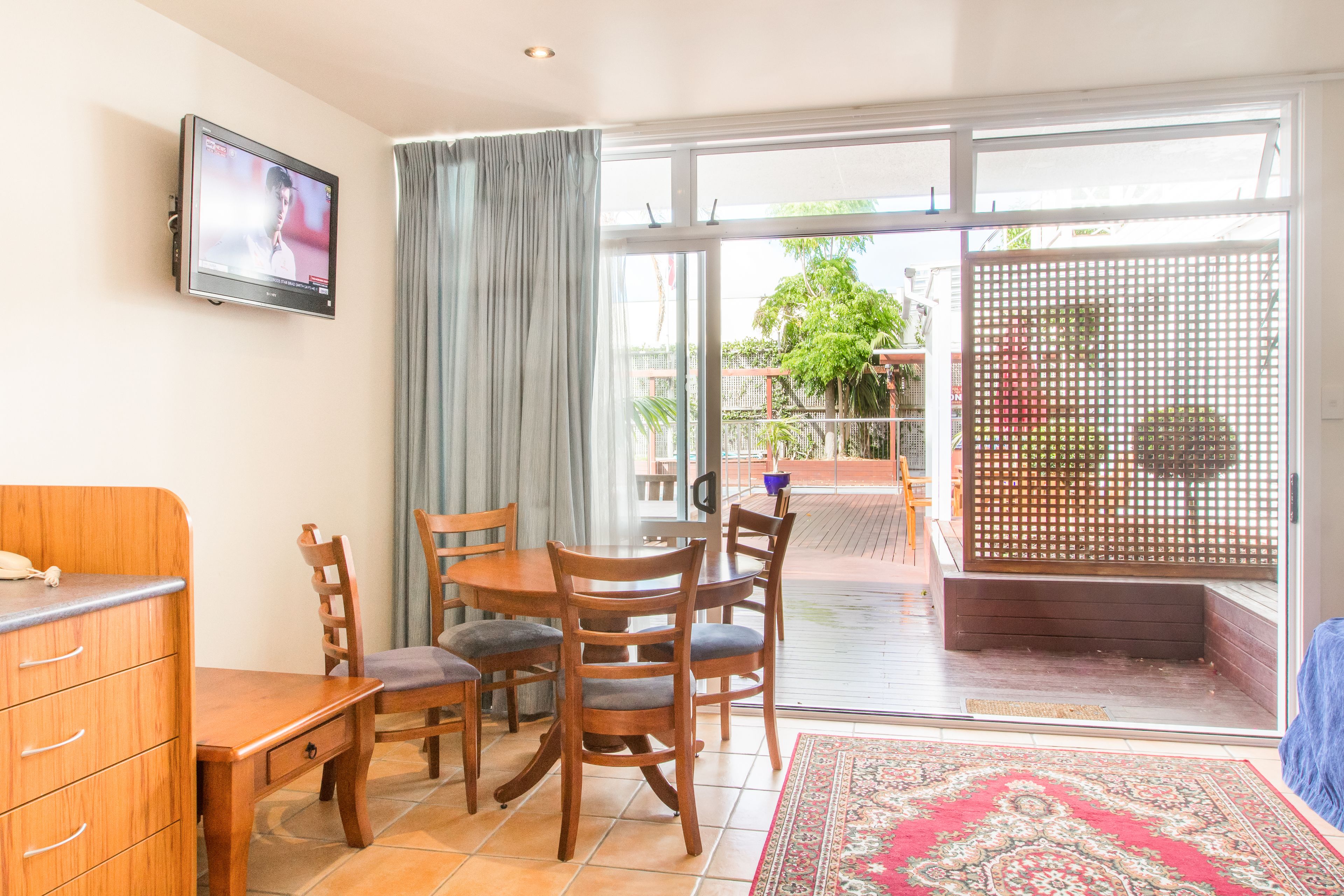 A room with a dining table, chairs, a TV mounted on the wall, a sliding glass door leading to an outdoor area, and a wooden cabinet.