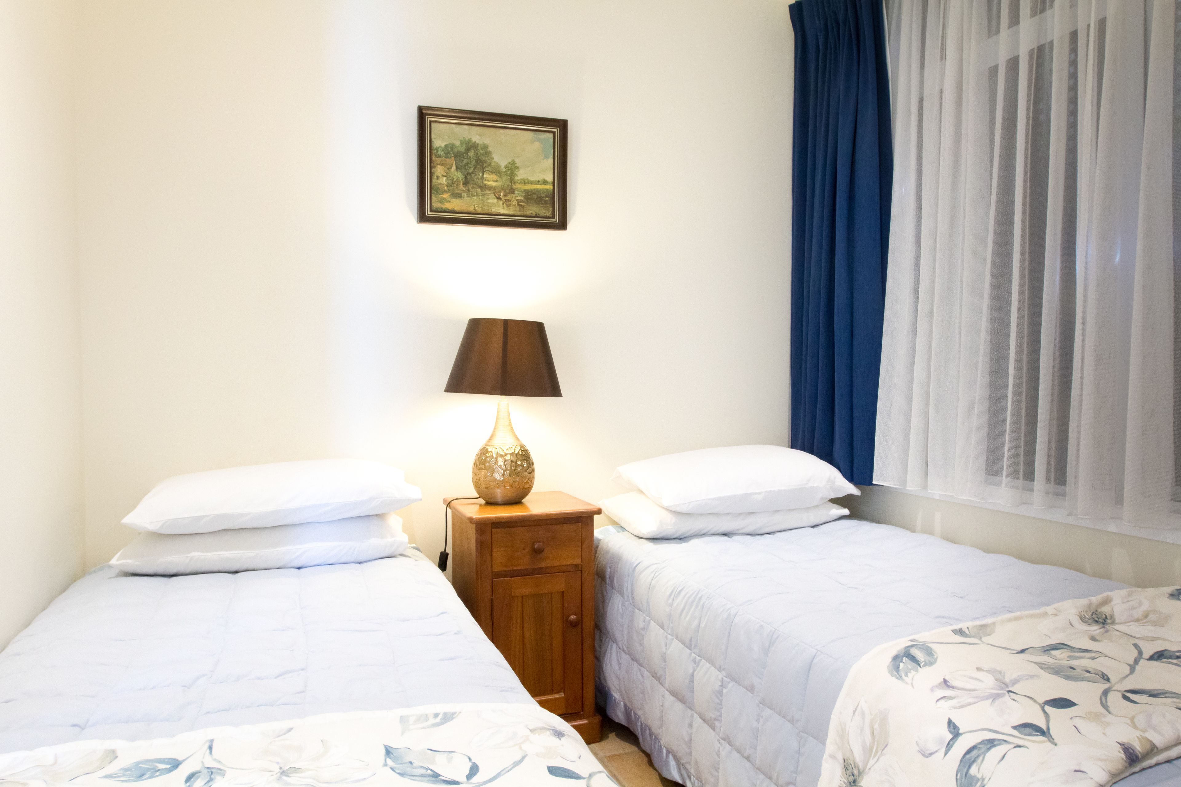 Un dormitorio con dos camas, una mesita de noche con lámpara, un cuadro enmarcado en la pared y una ventana con cortinas azules.