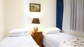 Un dormitorio con dos camas, una mesita de noche con lámpara, un cuadro enmarcado en la pared y una ventana con cortinas azules.