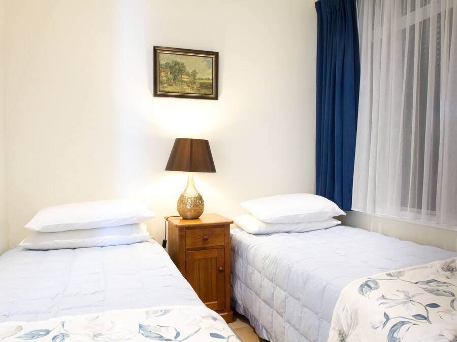 Un dormitorio con dos camas, una mesita de noche con lámpara, un cuadro enmarcado en la pared y una ventana con cortinas azules.