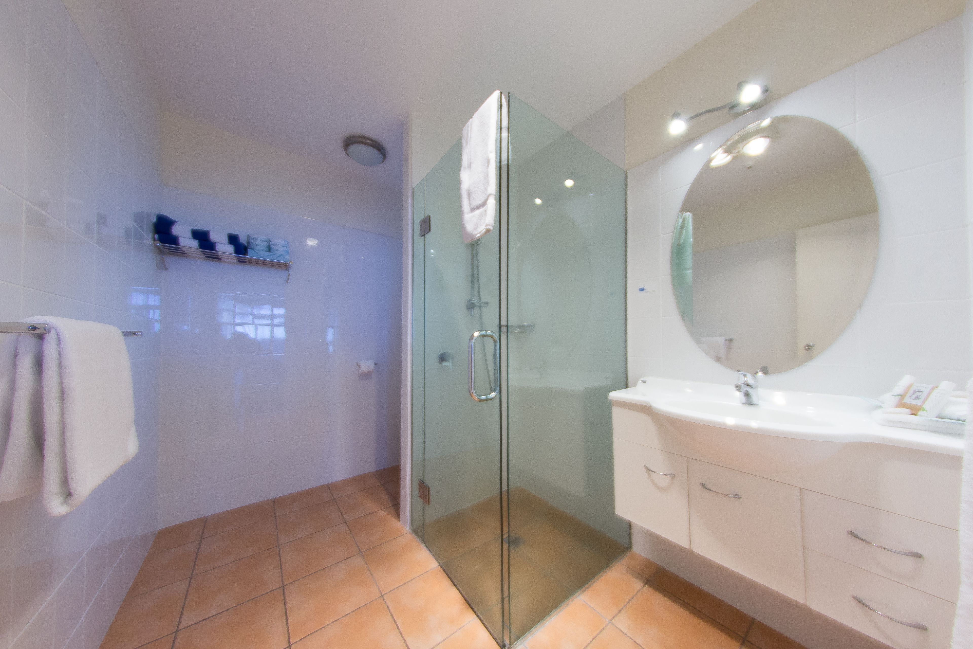 A modern bathroom with a glass-enclosed shower, a large round mirror, and a white vanity with drawers.