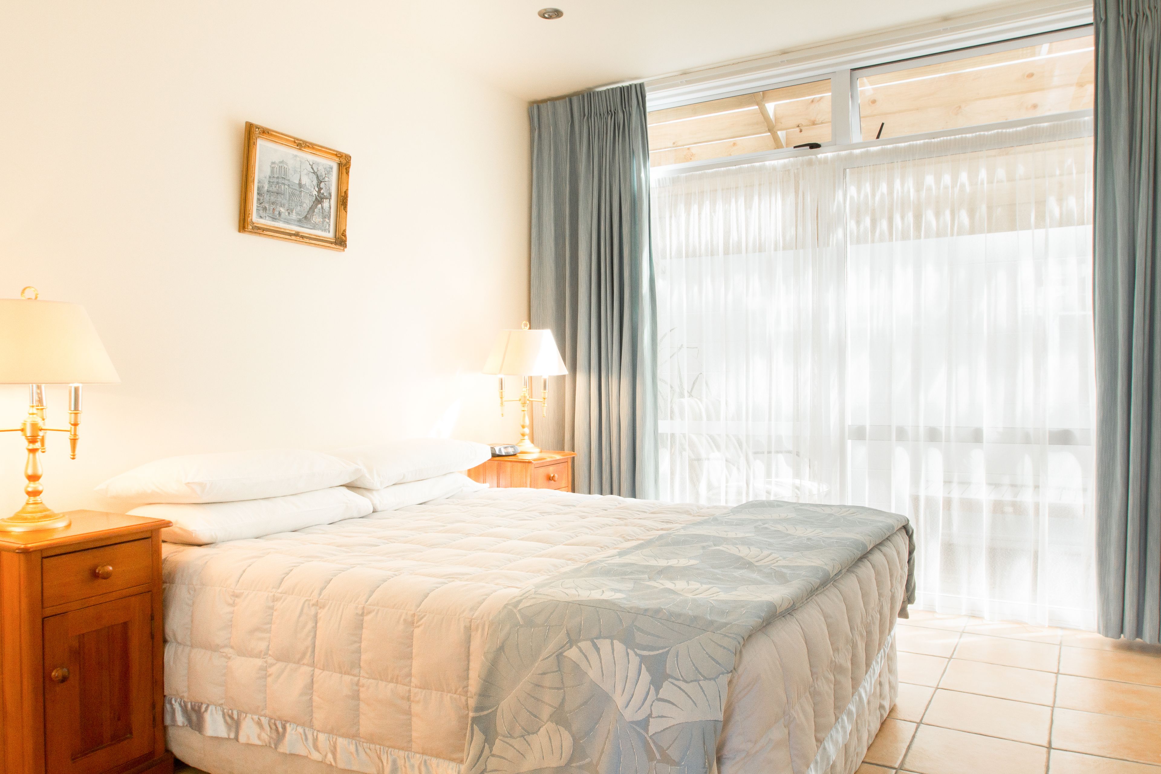 Une chambre à coucher avec un lit, deux tables de chevet avec des lampes, un tableau encadré au mur et une grande fenêtre avec des rideaux.